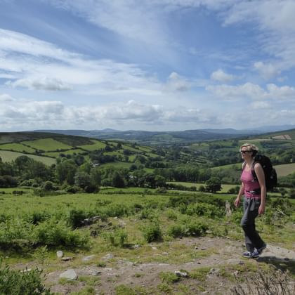 Vrouwelijke wandelaar met rugzak staat op rotsachtig terrein met uitzicht over groene heuvels en dalen van het Ierse platteland op de Wicklow Way.