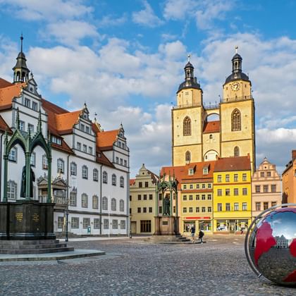 Historisch marktplein in Wittenberg met kleurrijke renaissancegebouwen, stadskerk met dubbele torens, sierfontein en rode bolsculptuur.
