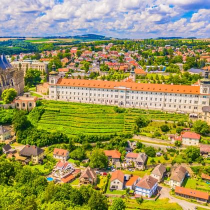 Luchtfoto van Kutná Hora met gotische kathedraal, terrasvormige wijngaarden en historische gebouwen met oranje daken te midden van groen landschap.