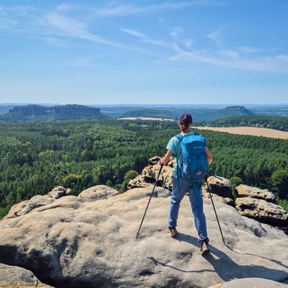 Wandelaar met blauwe rugzak en wandelstokken op rotsformatie bij Gohrisch, uitzicht over boslandschap met velden.