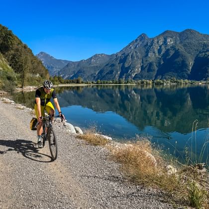 Fietser in geel shirt op grindpad langs Idromeer met bergen weerspiegeld in kalm water onder heldere blauwe lucht.