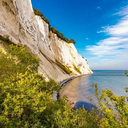 Dramatische witte krijtrotsen van Møns Klint die oprijzen vanaf de Oostzeekust, met groene vegetatie bovenop en smal strand eronder onder blauwe lucht.