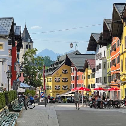 Kleurrijke gebouwen langs Kitzbühels voetgangerszone met buitenterrassen, kerktoren en Alpen op de achtergrond onder blauwe lucht.