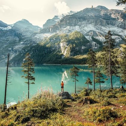 Wandelaar in rode jas staat op rotsuitstulping met uitzicht op turquoise Oeschinensee omringd door besneeuwde bergen en bos.