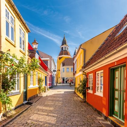 Geplaveide straat met kleurrijke huizen in geel, oranje en rood die leidt naar een klokkentoren in Faaborg, Denemarken.
