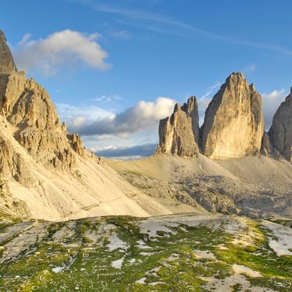 De iconische Drei Zinnen kalkstenen toppen in de Dolomieten onder blauwe hemel met witte wolken. Groene alpenweiden en rotsig terrein op voorgrond.