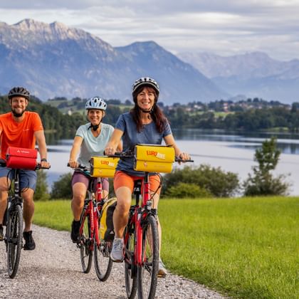 Drie fietsers op een grindpad langs het Riegsee-meer in Beieren, met groene weiden en dramatische Alpengebergte op de achtergrond.