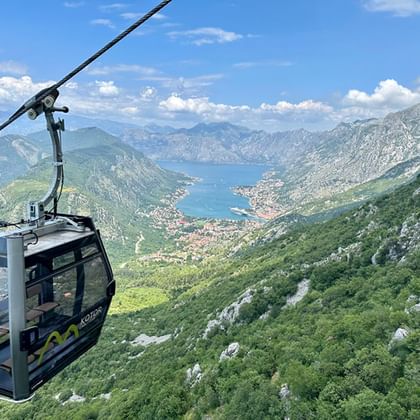 Zwarte kabelbaan cabine boven groene bergen met panoramisch uitzicht op de baai van Kotor en kustplaats onder blauwe lucht.