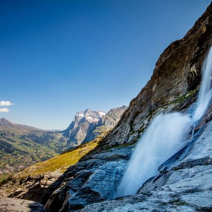 Sandbach waterval valt over rotsachtige klif in Berner Oberland met besneeuwde bergen en groene vallei op de achtergrond.