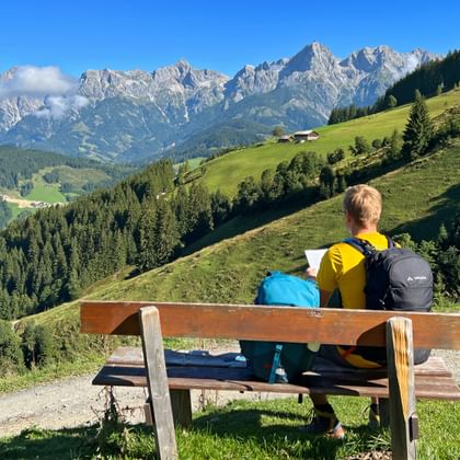 Twee wandelaars met rugzakken zitten op een houten bank met uitzicht op groene alpenweiden en de rotsachtige Kitzbüheler Alpen.
