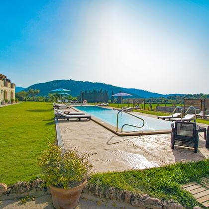 Rechthoekig hotelzwembad bij Monnaber Nou met ligstoelen, stenen gebouw, groen gazon en bergachtergrond onder blauwe lucht in Campanet.