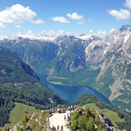 Wandelaars op berguitkijkpunt met uitzicht op Königssee meer omringd door met sneeuw bedekte Alpentoppen en beboste valleien.