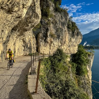 Fietser in geel shirt op Strada del Ponale, een schilderachtig klippad uitgehouwen in rotswand boven Gardameer met bergen op achtergrond.