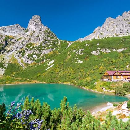 Turquoise bergmeer in het Tatra-gebergte met een gele hut met rood dak aan de oever, omringd door groene hellingen en rotsachtige toppen.
