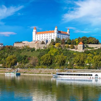 Wit riviercruiseschip op de Donau met kasteel Bratislava op beboste heuvel. Het kasteel heeft witte muren en rode daken onder blauwe lucht.