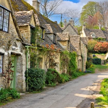 Historische stenen cottages van Arlington Row in Bibury met met mos bedekte daken en klimplanten, langs een glooiende dorpsweg in de Cotswolds.
