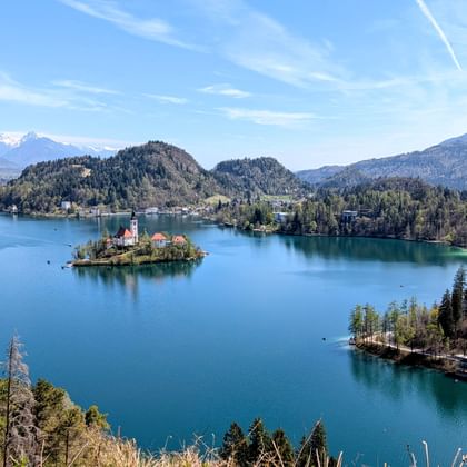 Panoramisch uitzicht op het meer van Bled in Slovenië met een klein bosrijk eiland met een kerk, omringd door bergen en blauw water onder heldere luchten.