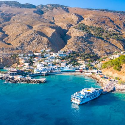 Luchtfoto van dorp Hora Sfakion aan Kreta's kust met witte gebouwen, turquoise baai, veerboot en bruine bergen onder blauwe lucht.