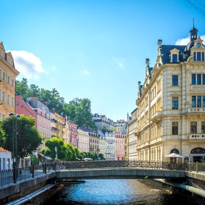 Kleurrijke historische gebouwen langs een rivier in Karlovy Vary, Tsjechië. Een brug kruist het water met een sierlijk geel gebouw rechts.