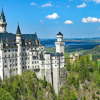 Kasteel Neuschwanstein op een rotsachtige heuvel omringd door groen bos en bergen onder een blauwe hemel met witte wolken.