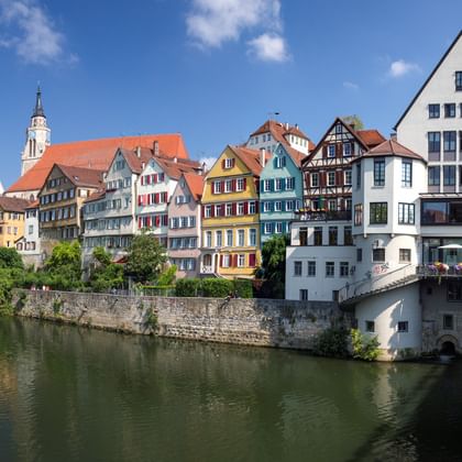Kleurrijke vakwerkhuizen langs de Neckar in Tübingen. Een stenen brug met bloembakken kruist het water, een kerktoren is zichtbaar.