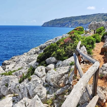 Houten wandelpad met leuningen langs rotsachtige kliffen in Marina Serra, Apulië. Blauwe Middellandse Zee en kustlijn zichtbaar.