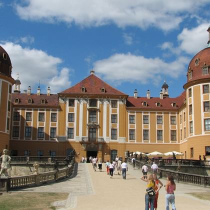 Kasteel Moritzburg met barokke torens Geel barok kasteel Moritzburg met rode koepeltorens en stenen brug. Bezoekers lopen over de dam onder blauwe hemel met wolken.
