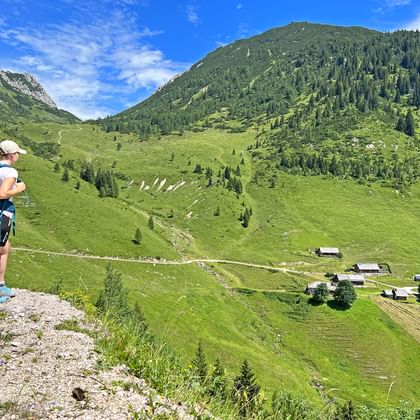 Vrouwelijke wandelaar met blauwe rugzak kijkt uit over de groene Rinnbergalm bergweidedal met traditionele alpenhutten en beboste bergen.