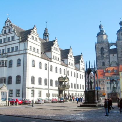 Marktplein in Lutherstadt Wittenberg met wit Renaissance stadhuis, kerk met dubbele torens, kleurrijke huizen en monument op kasseien.