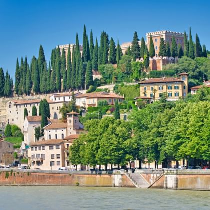 Kasteel Castel San Pietro op een heuvel in Verona, omringd door cipressen, met historische gebouwen eronder en de Adige op de voorgrond.