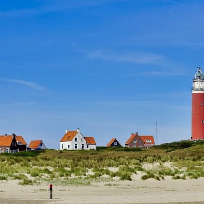 Vuurtoren Texel met dorpshuizen Rood-witte vuurtoren Texel staat hoog achter een rij traditionele Nederlandse huizen met oranje pannendaken, omgeven door duinen en gras.