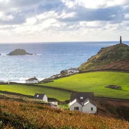 Glooiende groene velden en witte huisjes aan de kust van Cornwall. Een monument staat op een heuvel met uitzicht op de blauwe zee met rotseilanden.
