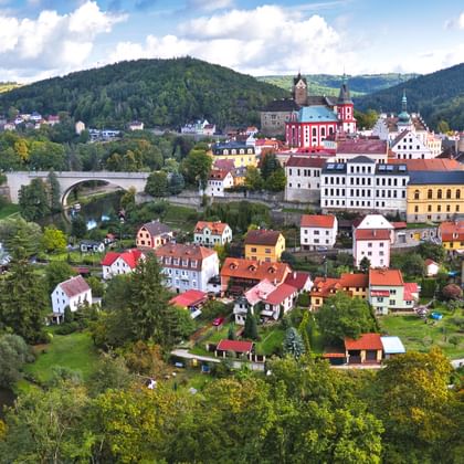 Panoramisch uitzicht op Loket, Tsjechië, met kleurrijke gebouwen, rode kerktoren, stenen brug over de Ohře, omringd door groene heuvels.