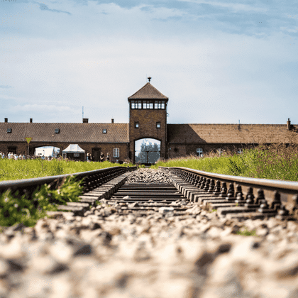 Spoorwegrails leiden naar de bakstenen poort met wachttoren van concentratiekamp Auschwitz. Groen gras groeit langs de rails onder een bewolkte hemel.