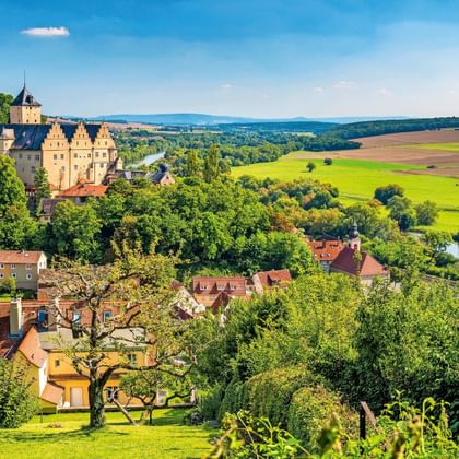 Kasteel Mainberg met uitzicht op een dorp met rode daken, omringd door groene bossen en velden langs de Main in Duitsland.