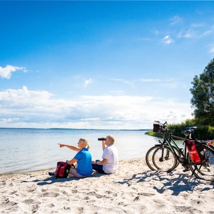 Twee fietsers zitten op een zandstrand met verrekijker, wijzend naar de zee. Twee toerfietsen met tassen staan nabij onder een blauwe lucht.