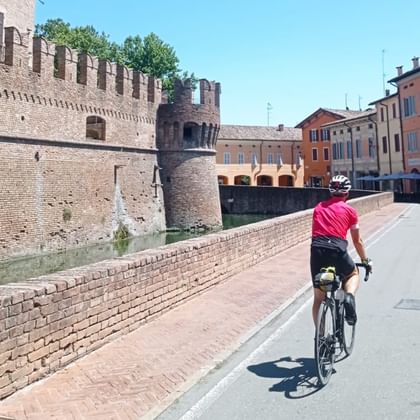 Fietser in roze shirt rijdt langs bakstenen vestingmuur met toren in Emilia, Italië. Kleurrijke historische gebouwen langs de straat.