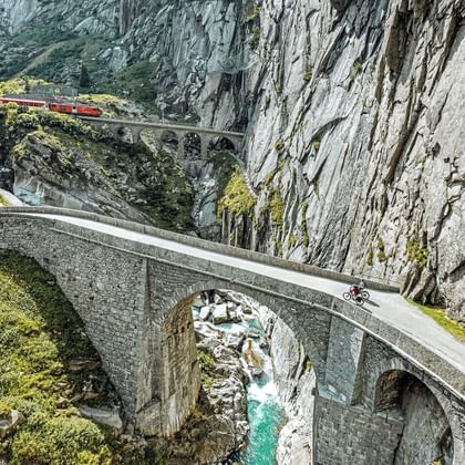 Luchtfoto van Duivelsbrug bij Andermatt met fietser op historische stenen boogbrug over turquoise rivier in rotskloof.
