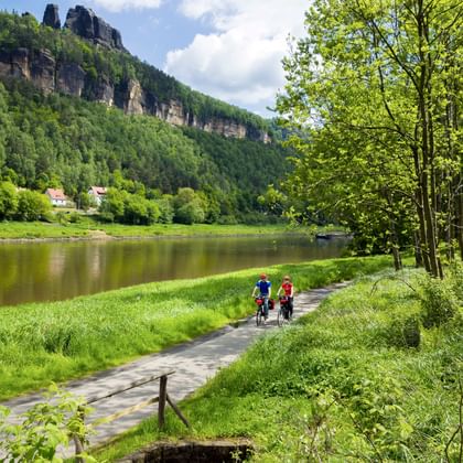 Twee fietsers op verharde weg bij Bad Schandau naast kalme rivier, met dramatische zandstenen kliffen en groen bos op achtergrond.