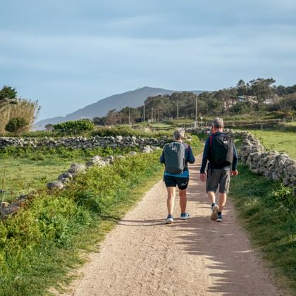 Twee wandelaars op een grindpad tussen stenen muren bij Oia. Groene velden en bergen zijn zichtbaar op de achtergrond onder een bewolkte hemel.