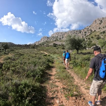 Drie wandelaars met rugzakken op een onverhard pad in Rhodos, Griekenland. Rotsachtige bergen en olijfbomen onder blauwe lucht met wolken.