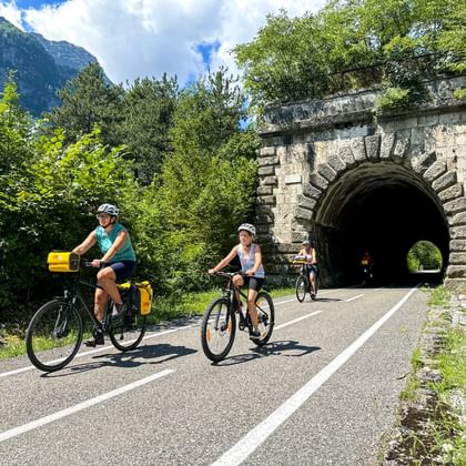 Fietsersgezin op Alpe Adria route bij historische stenen tunnel nabij Dogna, omringd door groene bergen en bomen.