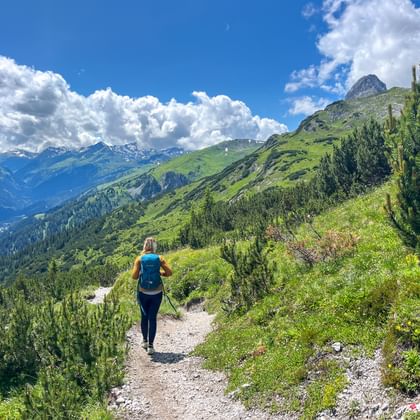 Vrouwelijke wandelaar met rugzak op bergpad door groen alpenlandschap met besneeuwde toppen en uitzicht op dal.