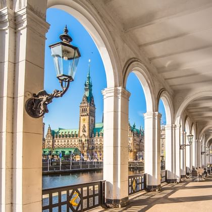 Zicht op Hamburgs historische stadhuis met groene koperen koepel door witte stenen bogen van een zuilengalerij. Traditionele straatlantaarn op voorgrond.