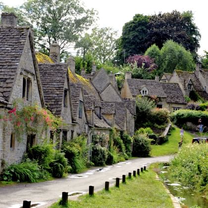Rij traditionele honingkleurige stenen huisjes in de Cotswolds met leien daken, tuinen en een beekje langs de weg.