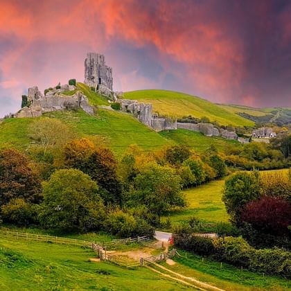 Ruïne van Corfe Castle op groene heuvel in Dorset onder dramatische oranje en paarse zonsonderganglucht, omringd door herfstbomen.
