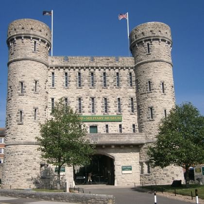 Stenen kasteelgebouw met twee ronde torens en kantelen in Dorchester. Het bord van het Keep Military Museum is zichtbaar boven de toegangsboog.