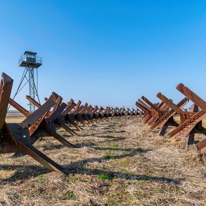 Rijen roestige metalen tankversperringen bij het IJzeren Gordijn Monument in Šatov, Tsjechië, met een wachttoren op de achtergrond onder blauwe lucht.