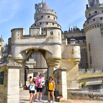 Drie bezoekers lopen door de stenen toegangspoort van Château de Pierrefonds. Het middeleeuwse kasteel heeft ronde torens met kantelen en een grote boogpoort.