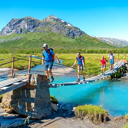 Groep wandelaars steekt houten Gjendebu-brug over turquoise rivier over in Jotunheimen Nationaal Park met besneeuwde bergen op achtergrond.